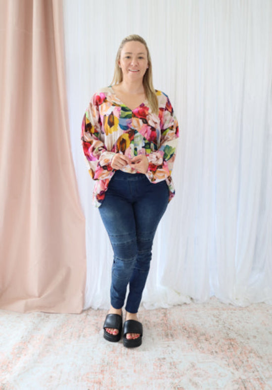 Woman wearing a colorful floral top and blue jeans standing against a white curtain.