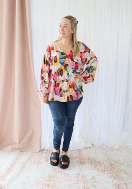 Woman wearing a colorful floral blouse and blue jeans against a white curtain background