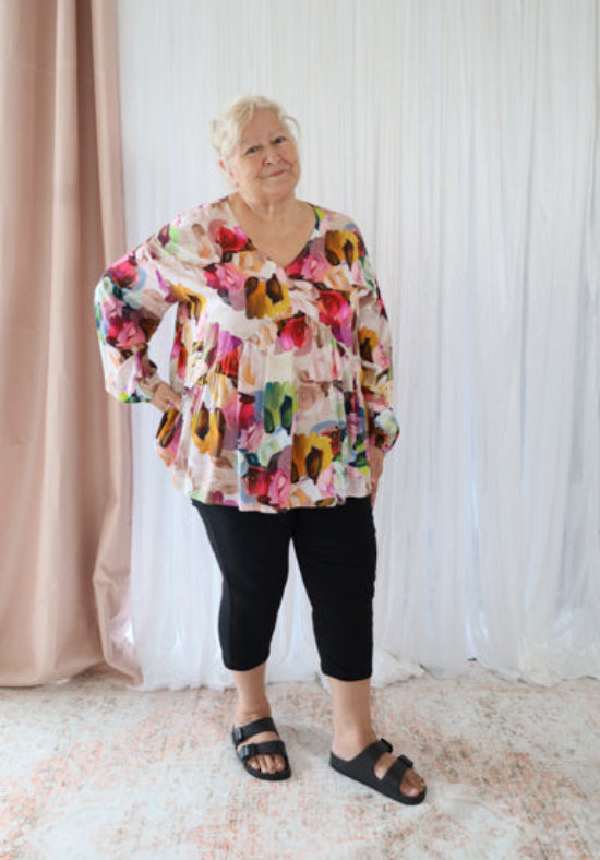 Woman wearing a colorful floral blouse and black pants standing in front of a white curtain.