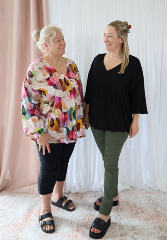 Two women standing side by side wearing colorful and black tops with green pants against a white curtain background.