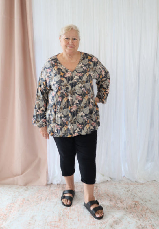 Person wearing a floral blouse and black pants standing against a white curtain.