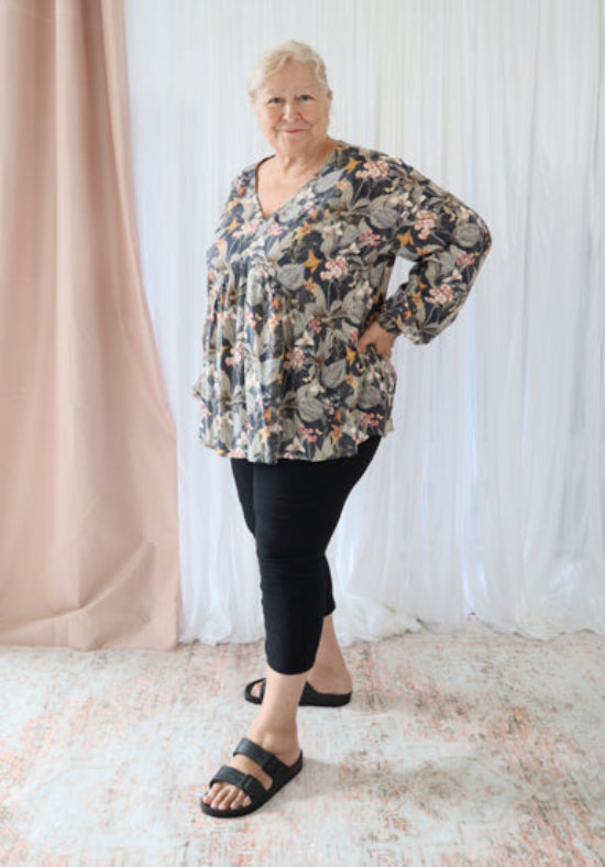 Woman wearing a floral blouse and black pants standing in front of light curtains.