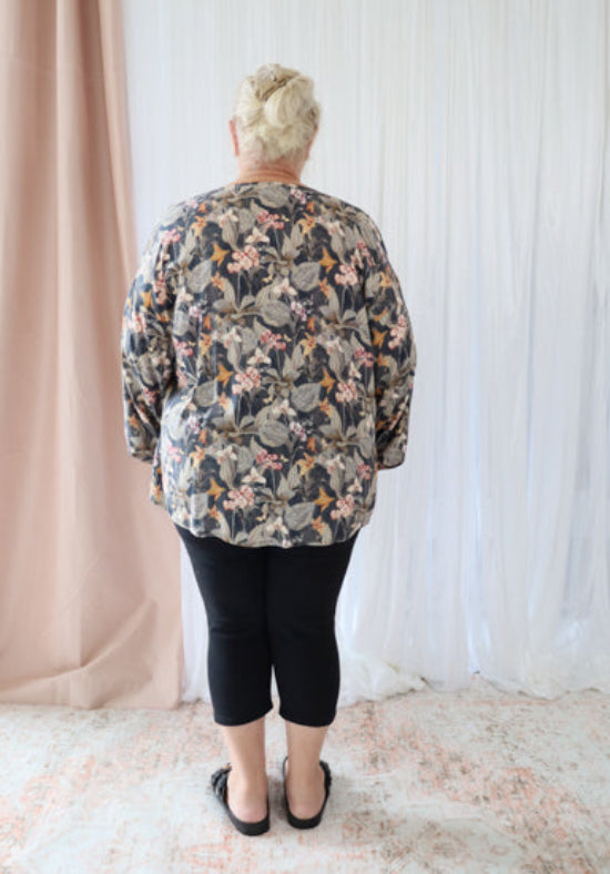Person wearing a floral blouse and black pants standing in front of a white curtain.