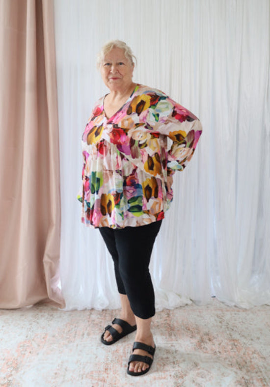 Woman wearing a colorful floral blouse and black pants standing against a white curtain.