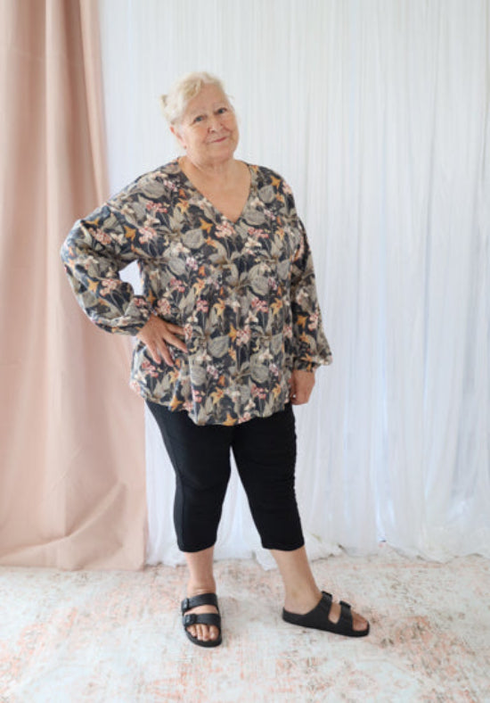 Woman wearing a floral blouse and black leggings standing against a white curtain.