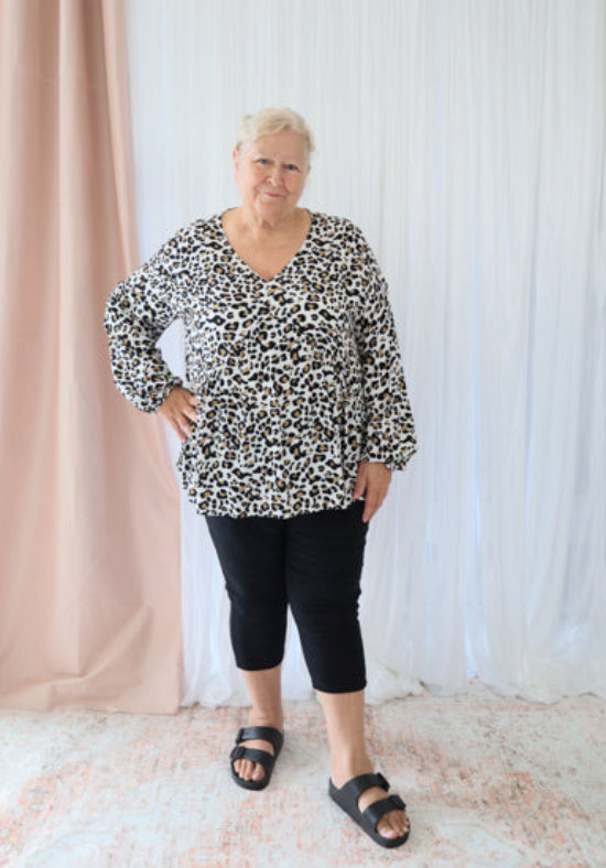Woman wearing a leopard print top and black pants standing against a white curtain.