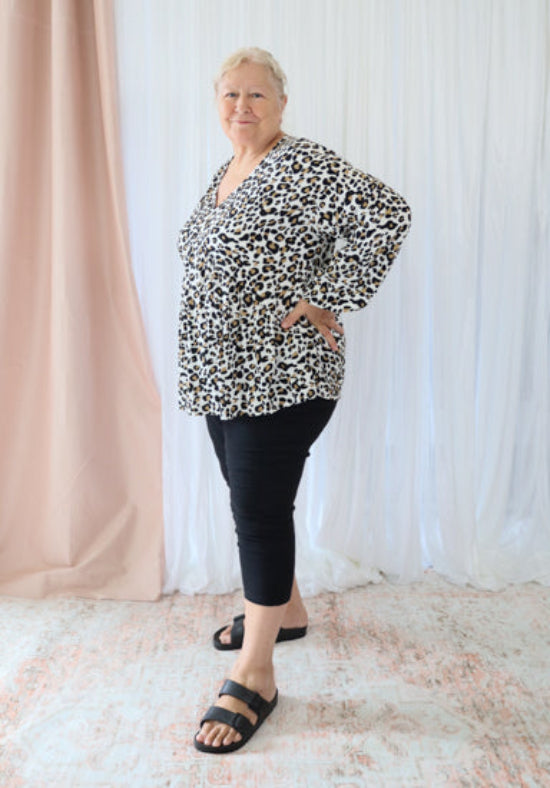 Woman wearing a leopard print top and black pants standing against a white curtain.