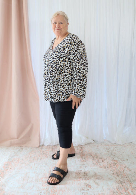 Woman wearing a leopard print top and black pants standing against a white curtain.
