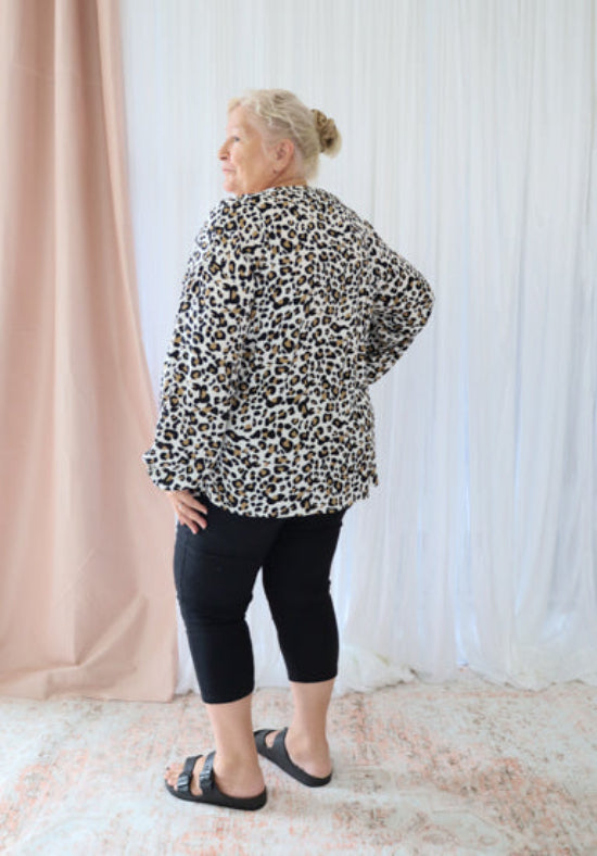 Person wearing a leopard print jacket and black pants standing against a light curtain background