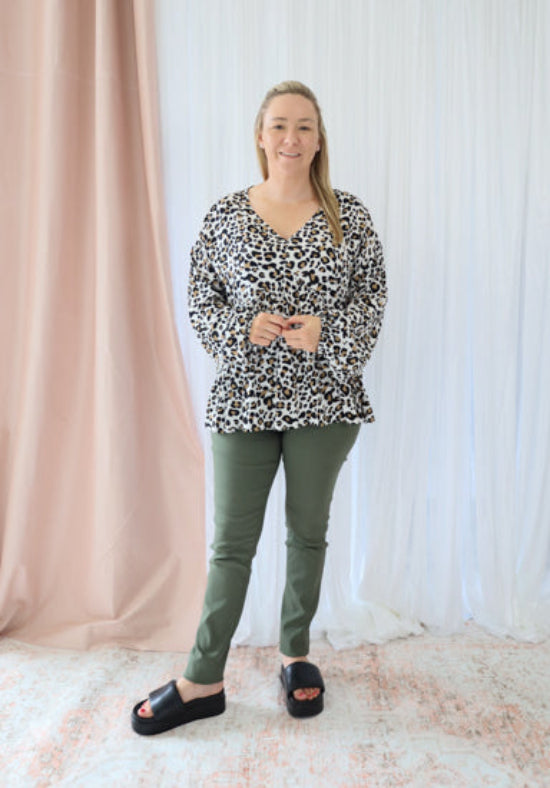 Woman wearing a leopard print top and green pants standing in front of a white curtain.