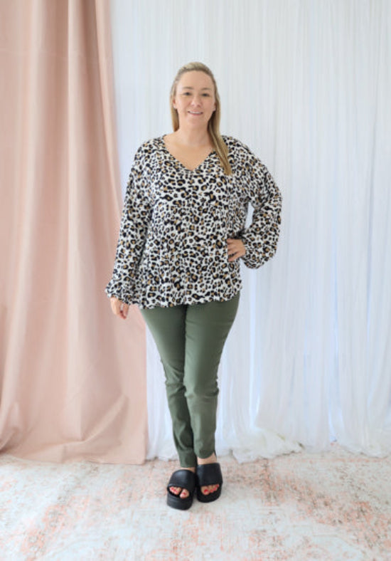 Woman wearing a leopard print top and green pants standing in front of a white curtain.