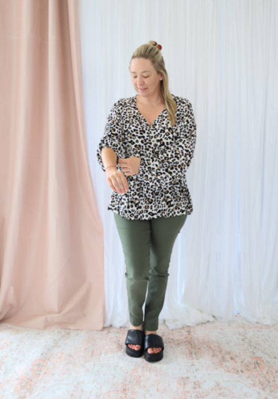 Woman wearing a leopard print top and green pants standing in front of a white curtain.