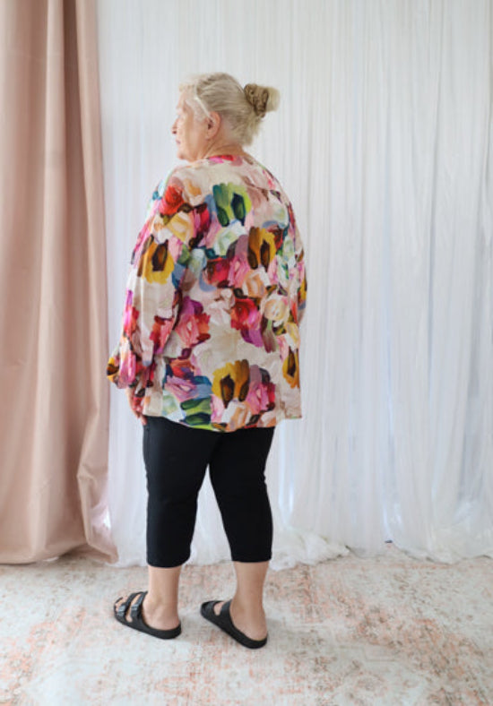Person wearing a colorful floral jacket standing in a room with light-colored walls and a pink curtain.