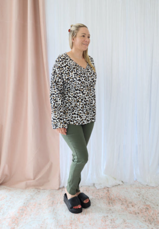 Woman wearing a leopard print top and green pants standing in front of a white curtain.