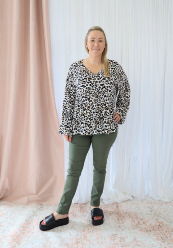 Woman wearing a leopard print top and green pants standing in front of a white curtain.