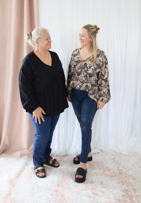 Two women standing side by side against a white curtain with a pink curtain in the background.