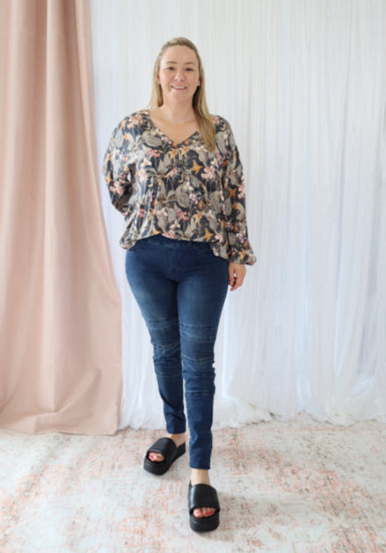 Woman wearing a floral blouse and blue jeans standing against a white curtain.