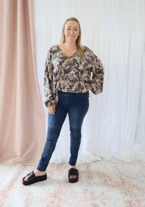 Woman wearing a floral blouse and blue jeans standing against a white curtain background