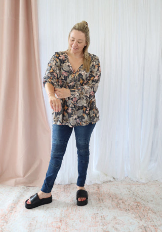 Woman wearing a floral blouse and blue jeans standing against a white curtain background