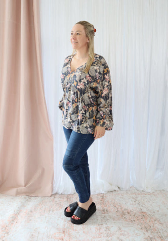 Woman wearing a floral blouse and blue jeans standing against a white curtain.