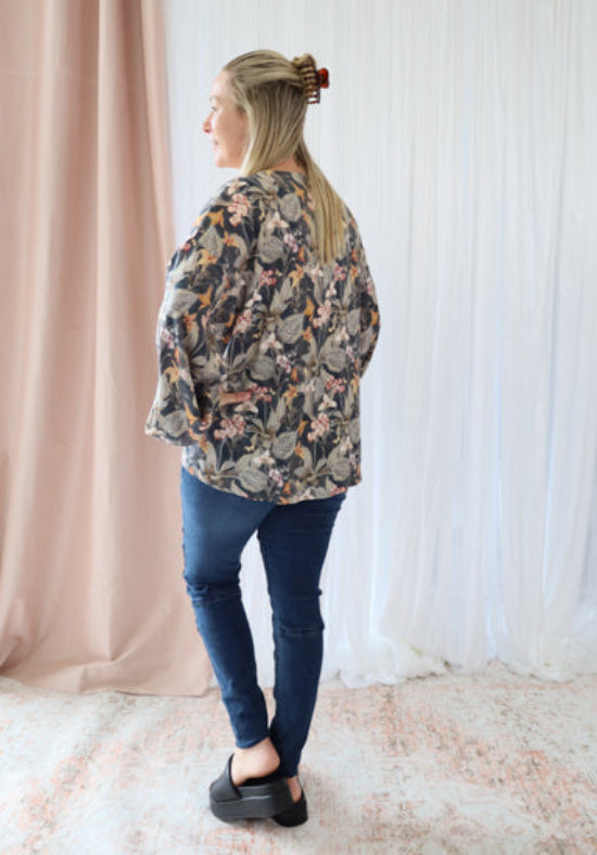 Woman wearing a floral jacket and blue jeans standing against a white curtain.