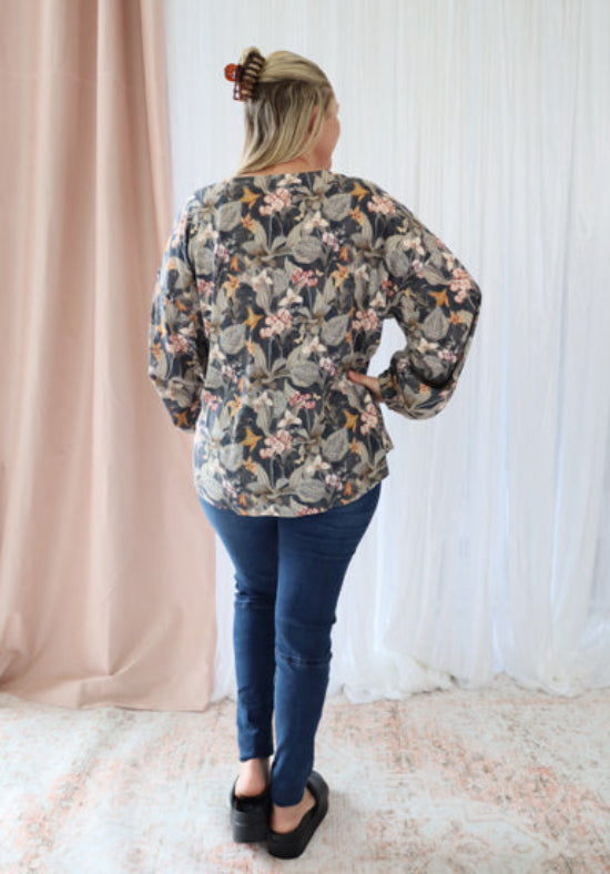 Person wearing a floral blouse and blue jeans standing in front of a white curtain.