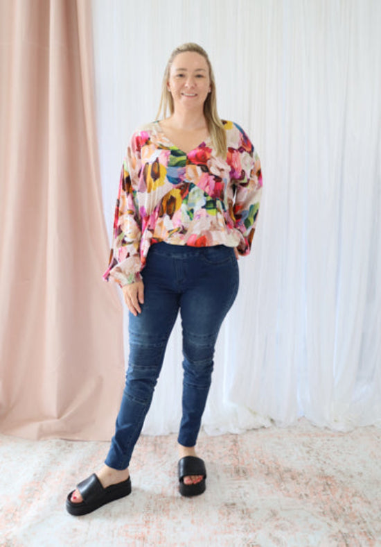 Woman wearing a colorful floral blouse and blue jeans standing against a white curtain background