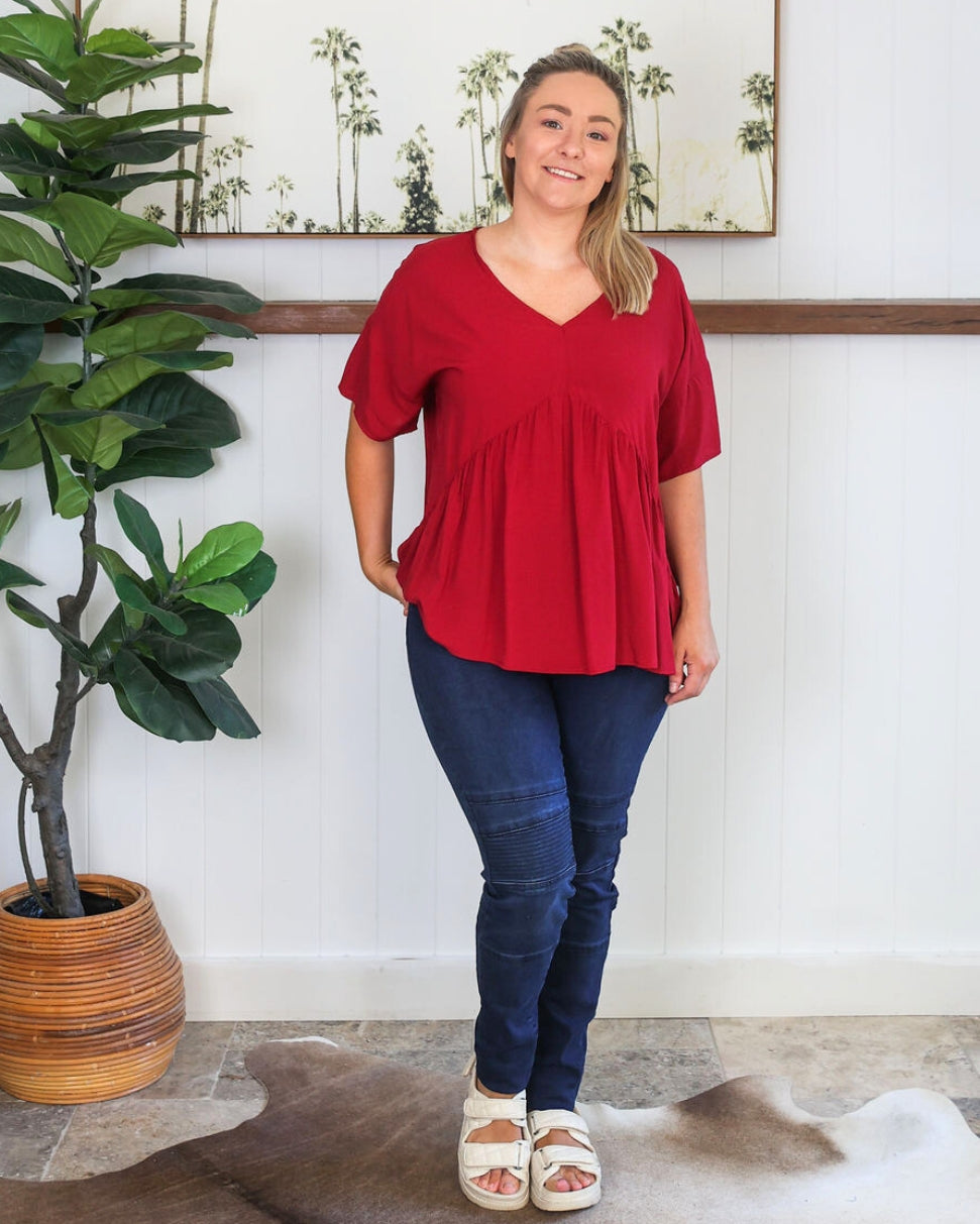 Woman wearing a red top and blue jeans standing indoors with a plant and rug in the background