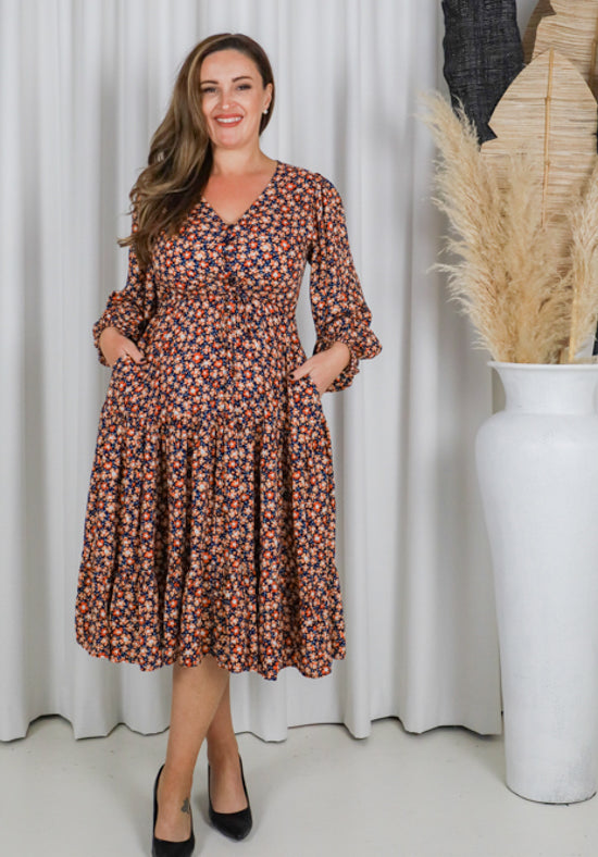 Woman wearing a floral dress standing in front of a white curtain with a vase of dried plants.