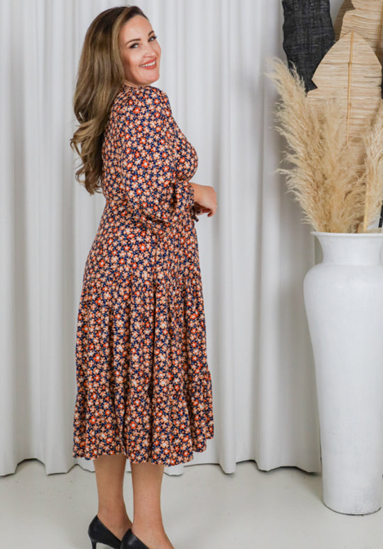 Woman in a floral dress standing next to a white curtain and decorative vase.