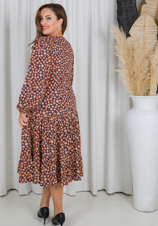 Woman wearing a floral dress standing in front of white curtains with a vase of dried plants.