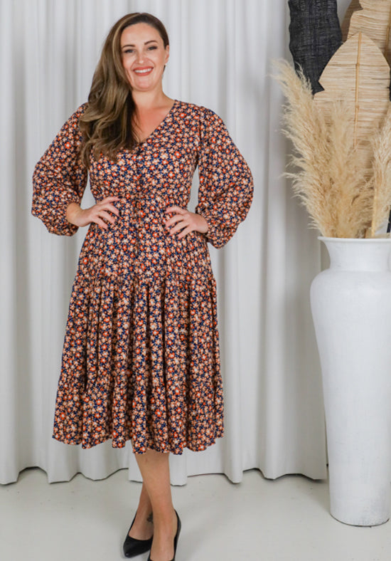 Woman wearing a floral dress standing in front of a white curtain with a vase of dried plants.