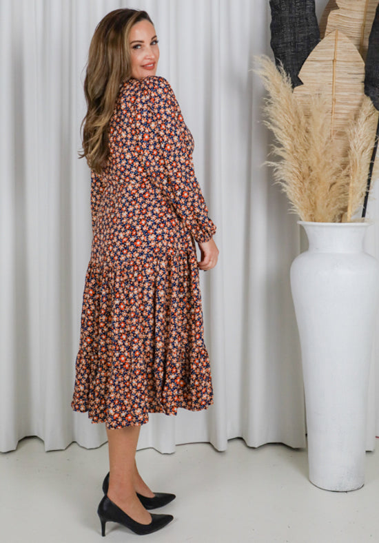Woman wearing a floral dress standing in front of white curtains with a vase of dried plants.