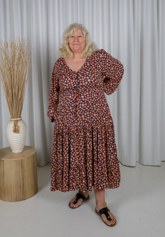 Woman wearing a floral dress standing in front of a white curtain with a vase on a wooden table.