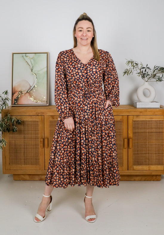 Woman wearing a floral dress standing in a room with wooden furniture and plants.