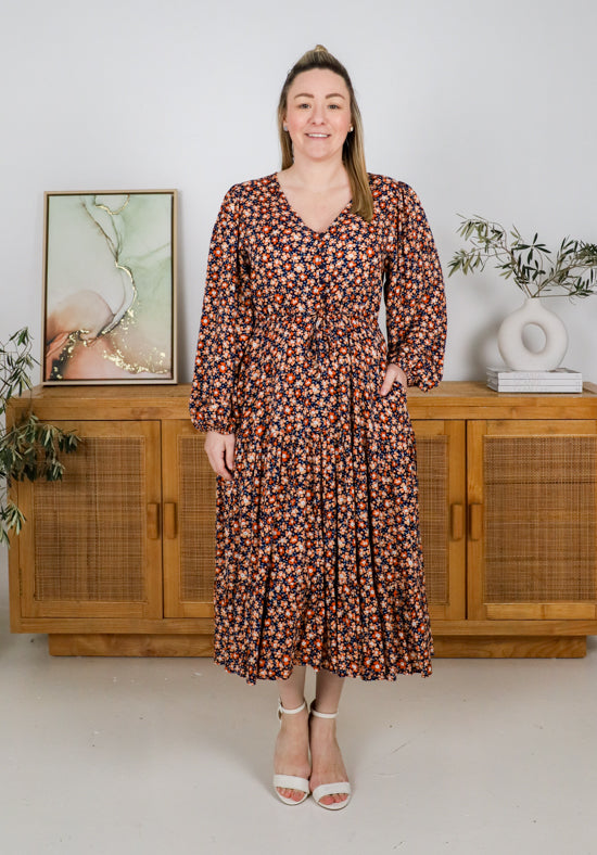 Woman wearing a floral dress standing in a room with wooden furniture and plants.