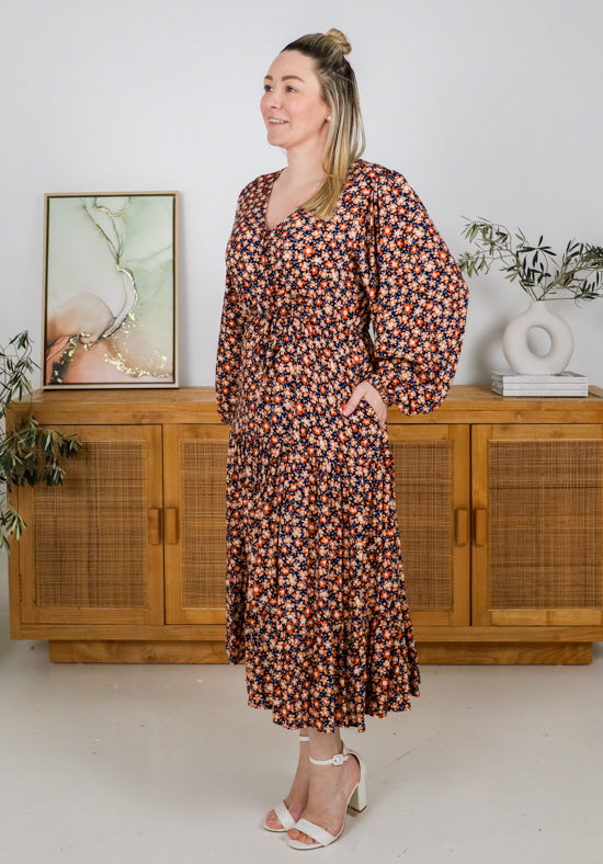 Woman wearing a floral dress standing in a room with wooden cabinets and plants.
