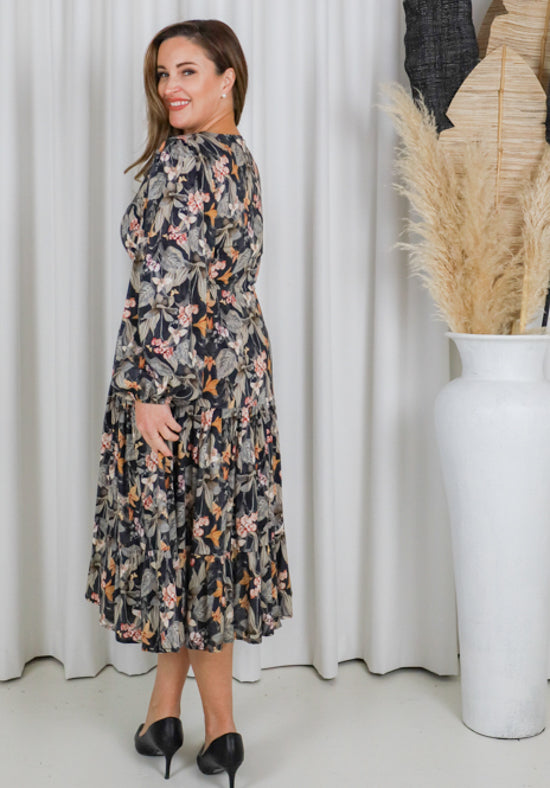 Woman wearing a floral dress standing in a room with wooden cabinets and plants.