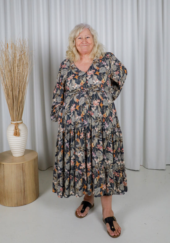 Woman wearing a floral dress standing in front of a white curtain with a vase of dried plants on a wooden table.