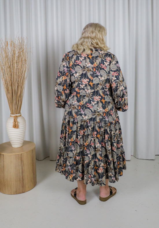 Woman wearing a floral dress standing in front of a white curtain with a vase of dried plants on a wooden table.