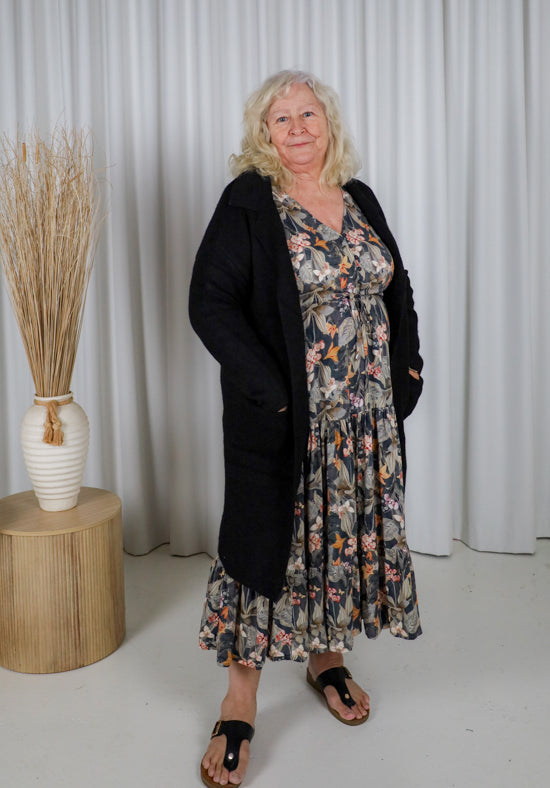 Woman wearing a floral dress and black cardigan standing in front of a white curtain.