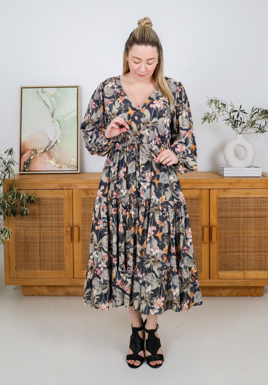 Woman wearing a floral dress standing in a room with wooden furniture and plants.