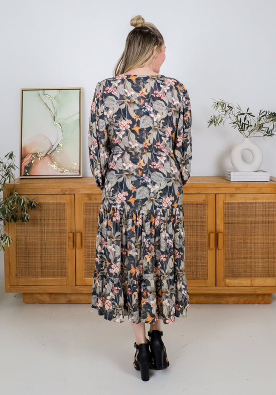 Woman wearing a floral dress standing in a room with wooden cabinets and plants.