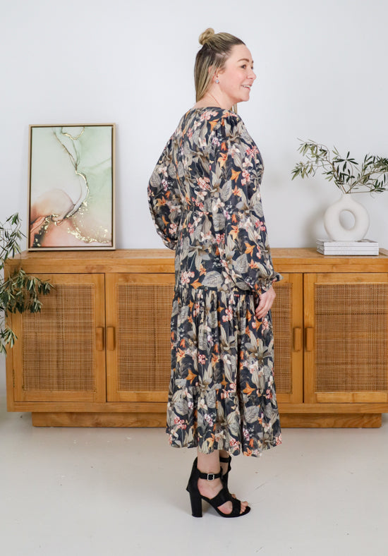 Woman wearing a floral dress standing in a room with wooden furniture and plants.