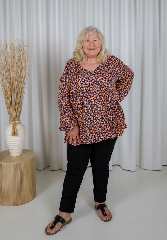Woman wearing a patterned blouse and black pants standing in front of white curtains.