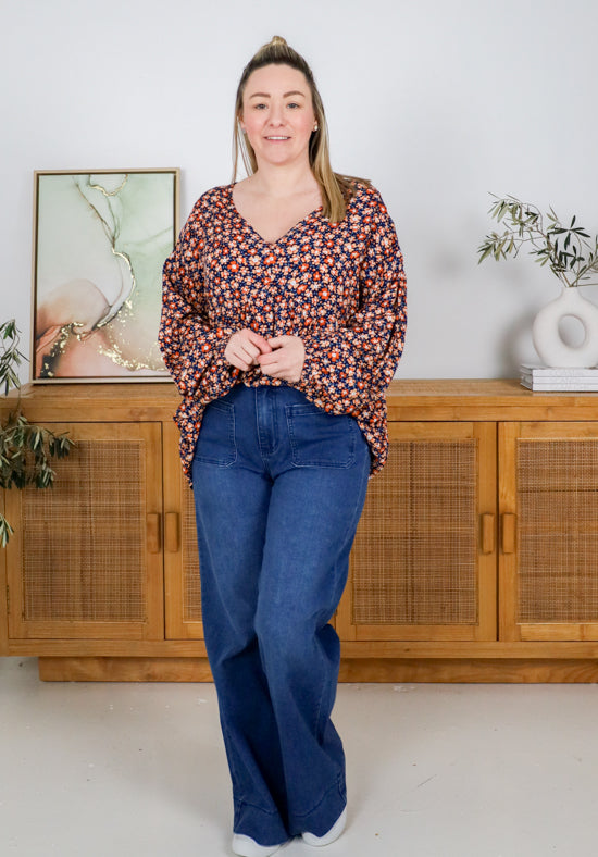 Woman wearing a patterned blouse and blue jeans standing in a room with wooden furniture and decor.