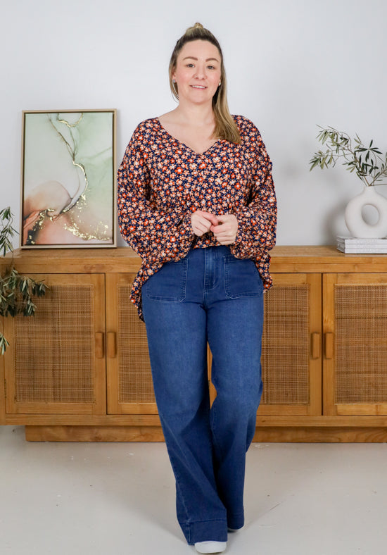 Woman wearing a floral blouse and blue jeans standing in a room with wooden furniture and plants.