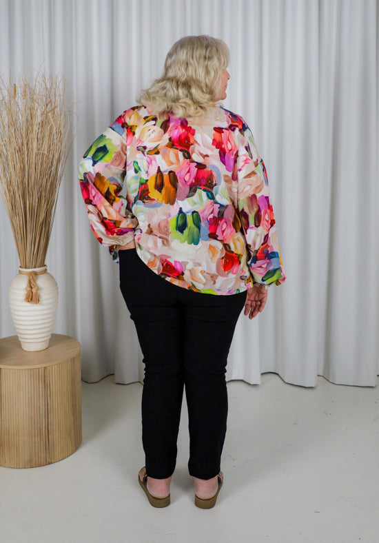 Woman wearing a colorful floral jacket standing against a white curtain.