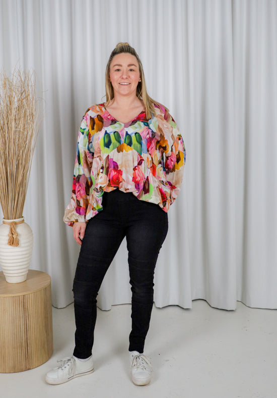 Person wearing a colorful floral blouse and black pants standing in front of a white curtain.
