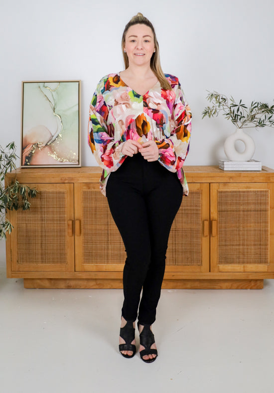Woman wearing a colorful floral blouse and black pants standing in a room with wooden furniture and plants.